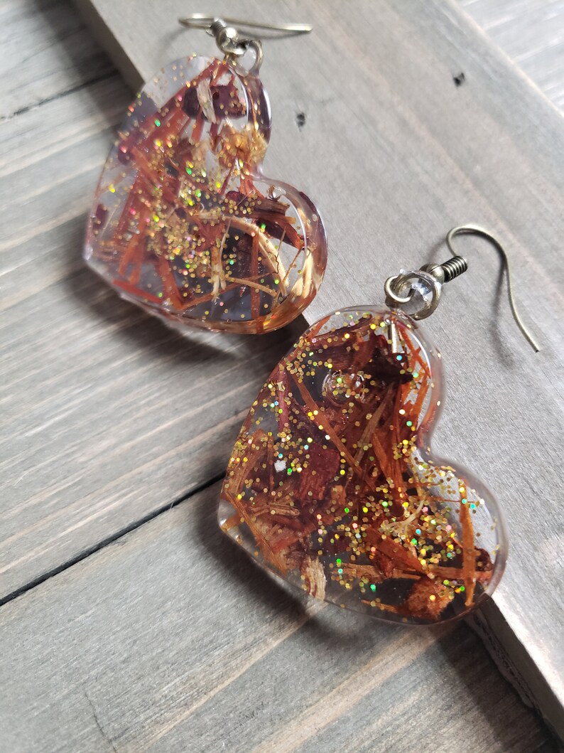 May include: A pair of heart-shaped resin earrings with gold glitter and brown wood-like inclusions. The earrings have silver hooks.