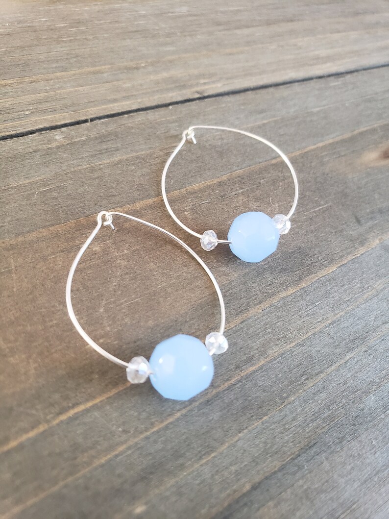 Puede incluir: Un par de pendientes de aro de plata con una piedra preciosa azul claro y detalles de cristal transparente.
