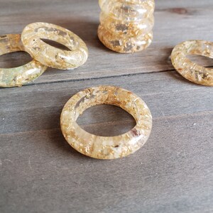 Op de afbeelding: Vijf transparante servetringen van hars met ingebedde gouden vlokken. De ringen zijn gestapeld op een houten oppervlak.