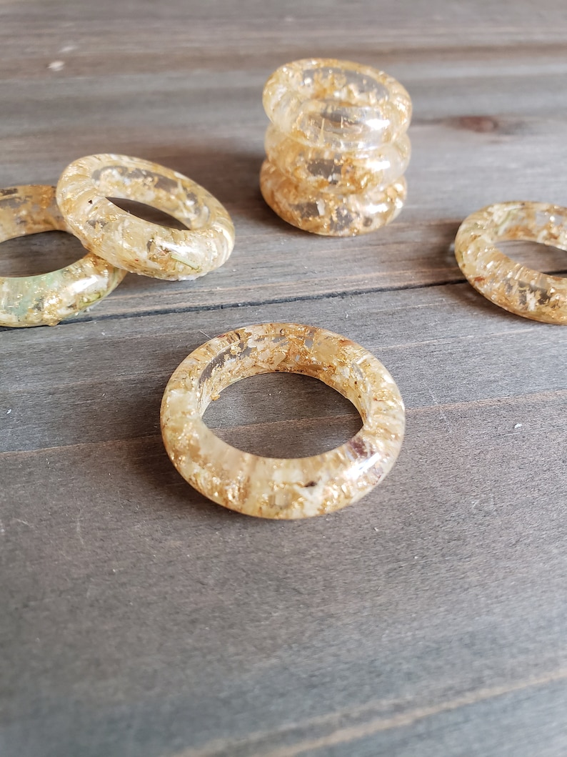 Op de afbeelding: Vijf transparante servetringen van hars met ingebedde gouden vlokken. De ringen zijn gestapeld op een houten oppervlak.