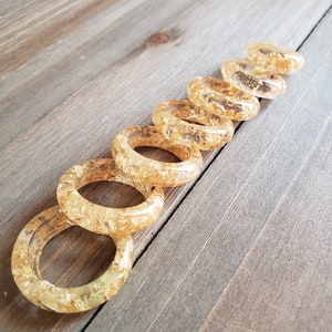 Op de afbeelding: Een set van acht gouden harsringen met ingebedde gouden vlokken. De ringen zijn in een lijn op een houten oppervlak gerangschikt, met de grootste ring bovenaan en de kleinste ring onderaan.