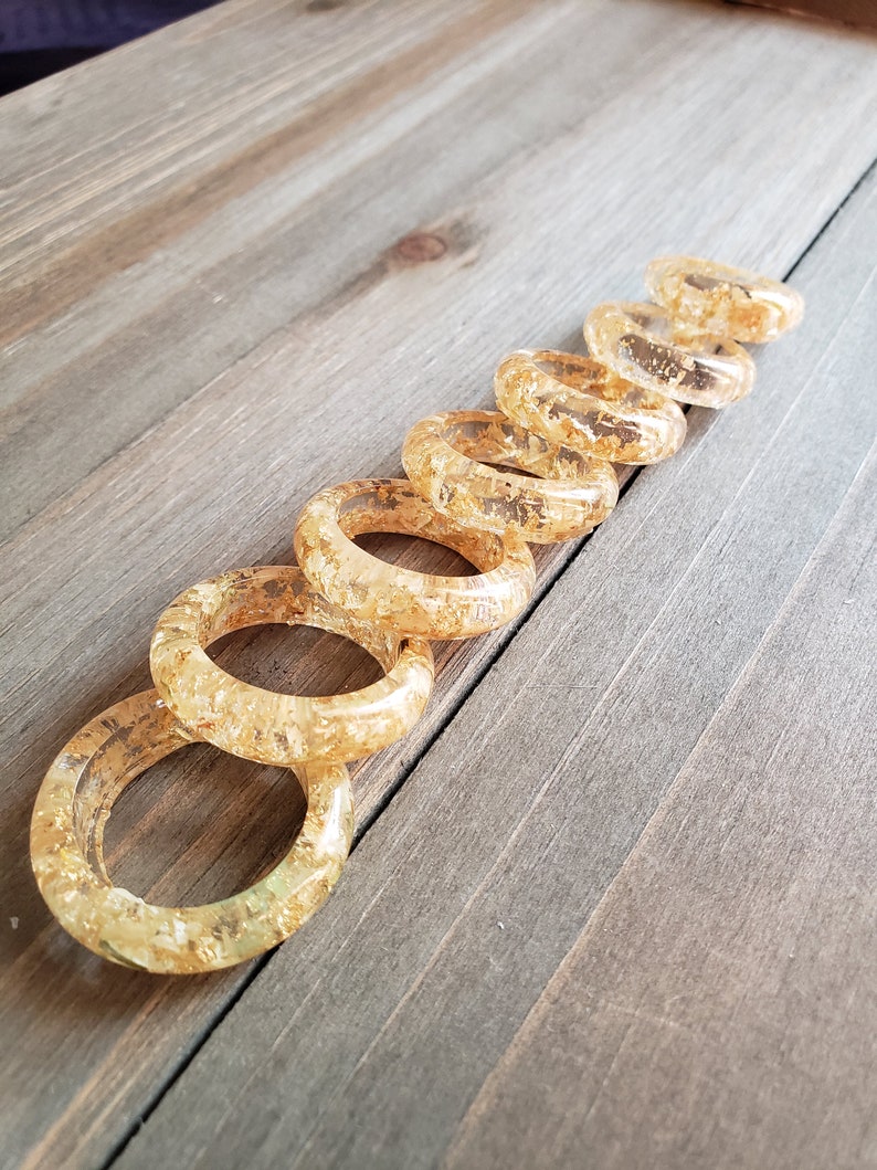 Op de afbeelding: Een set van acht gouden harsringen met ingebedde gouden vlokken. De ringen zijn in een lijn op een houten oppervlak gerangschikt, met de grootste ring bovenaan en de kleinste ring onderaan.