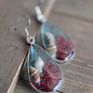 Puede incluir: Pendientes en forma de lágrima con marco plateado. Cada pendiente presenta una concha marina en miniatura, coral rojo y un fondo azul claro en resina. Los pendientes cuelgan de ganchos plateados.