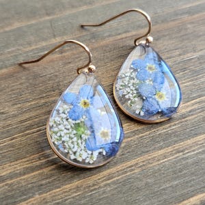 Puede incluir: Un par de pendientes en forma de lágrima con ganchos dorados. Cada pendiente presenta una lágrima de resina transparente con flores azules y blancas prensadas en su interior.