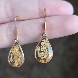 May include: A pair of teardrop-shaped earrings with gold-colored metal frames. Each earring features a clear resin enclosure containing dried, light-yellow flowers. The earrings have gold-colored fish hook ear wires.