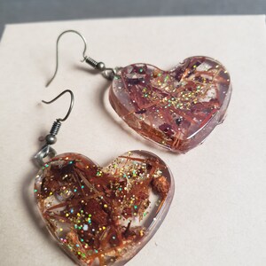 May include: A pair of heart-shaped earrings with brown and gold glitter embedded in clear resin. The earrings have silver hooks.