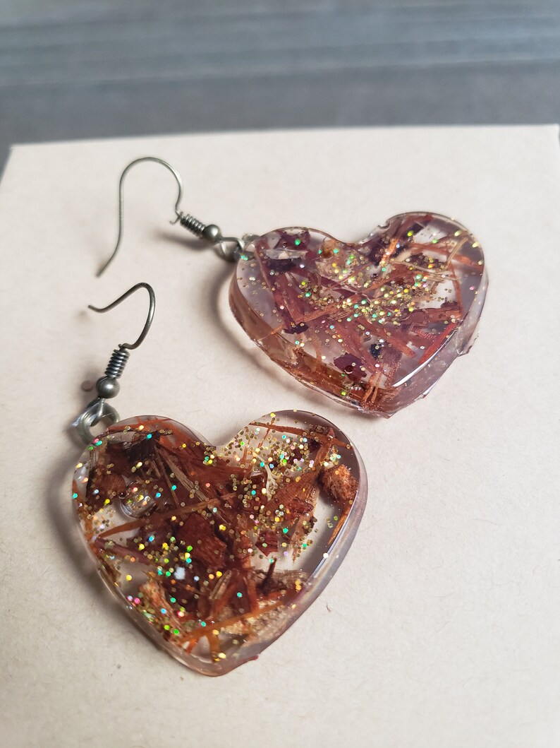 May include: A pair of heart-shaped earrings with brown and gold glitter embedded in clear resin. The earrings have silver hooks.