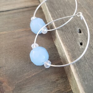 Puede incluir: Un par de pendientes de aro de plata con cuentas de piedras preciosas de color azul claro. Los pendientes est&aacute;n sobre una superficie de madera.