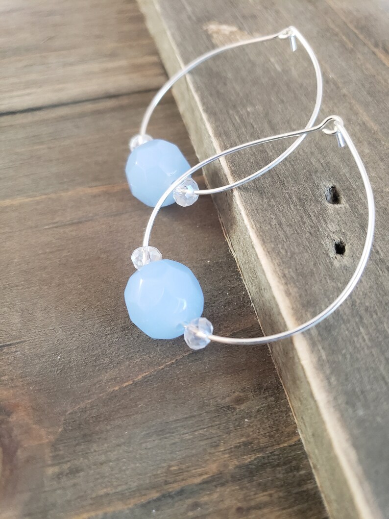 Puede incluir: Un par de pendientes de aro de plata con cuentas de piedras preciosas de color azul claro. Los pendientes est&aacute;n sobre una superficie de madera.