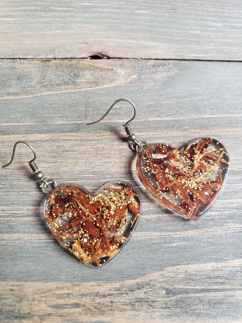 May include: A pair of heart-shaped resin earrings with gold glitter and brown wood-like pieces embedded inside. The earrings have bronze-colored hooks.