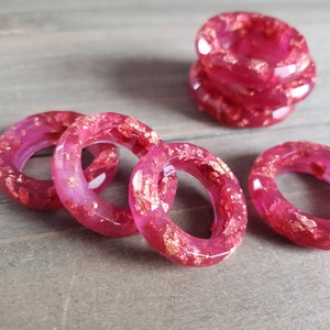 May include: Four pink resin rings with gold flecks. The rings are stacked on a wooden surface.