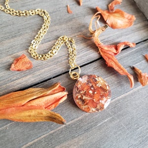 May include: A gold chain necklace with a round pendant featuring pressed orange flower petals encased in clear resin.