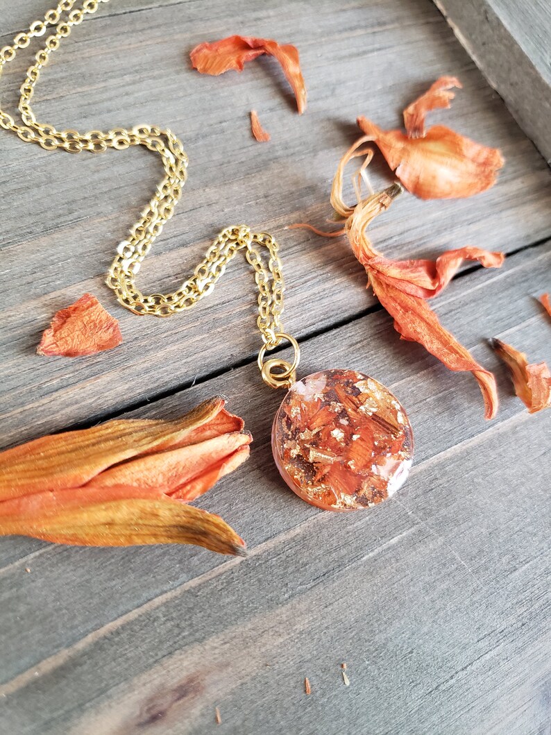 May include: A gold chain necklace with a round pendant featuring pressed orange flower petals encased in clear resin.