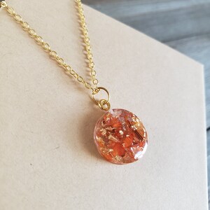 May include: A gold chain necklace with a round pendant featuring a clear resin filled with orange and gold flakes.