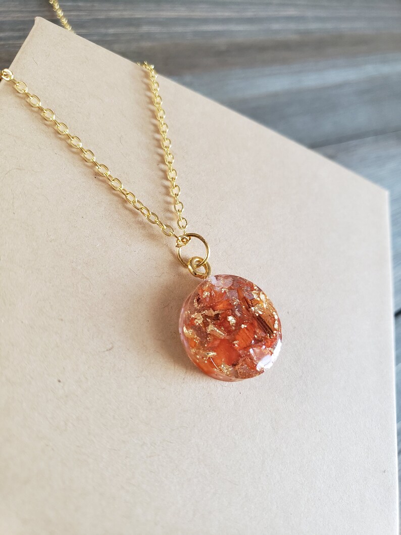 May include: A gold chain necklace with a round pendant featuring a clear resin filled with orange and gold flakes.