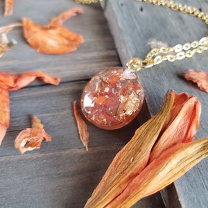 May include: A gold chain necklace with a round pendant featuring dried orange flower petals and gold leaf encased in clear resin.