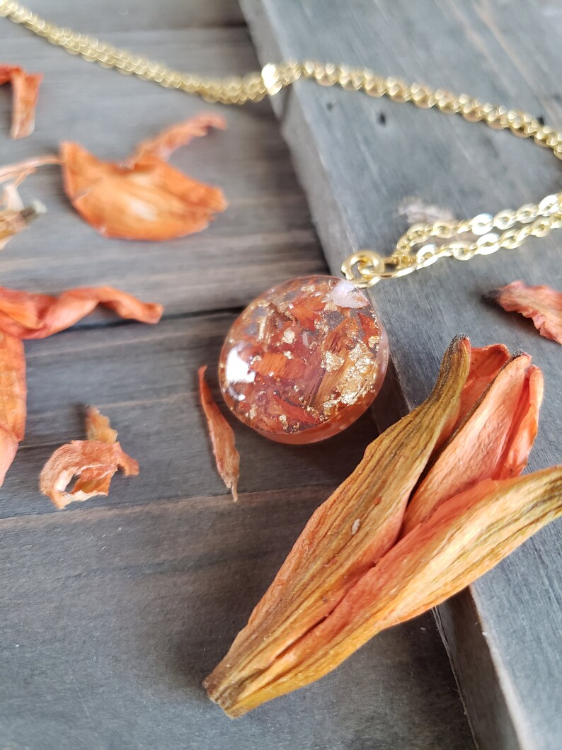 May include: A gold chain necklace with a round pendant featuring dried orange flower petals and gold leaf encased in clear resin.