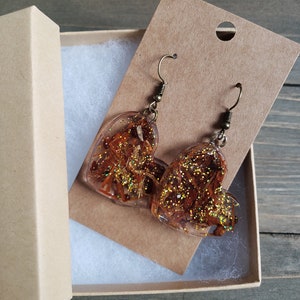 May include: A pair of heart-shaped resin earrings with gold glitter and dried flowers. The earrings are on a brown card with a hole for hanging.