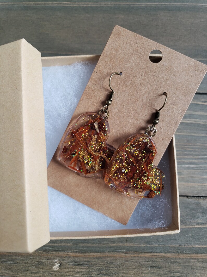 May include: A pair of heart-shaped resin earrings with gold glitter and dried flowers. The earrings are on a brown card with a hole for hanging.