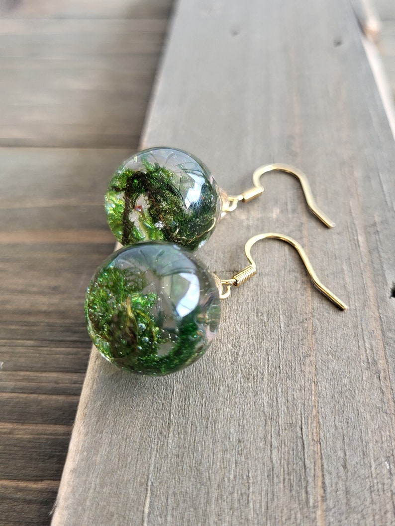 May include: A pair of gold earrings with clear glass orbs containing green moss and glitter.