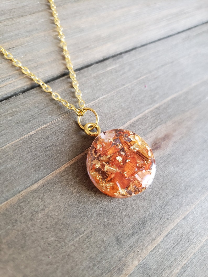 May include: A gold chain necklace with a round pendant featuring a resin design with orange and gold flakes.