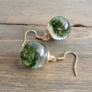 May include: A pair of gold earrings with clear glass orbs containing preserved green moss. The orbs are suspended from gold hooks.