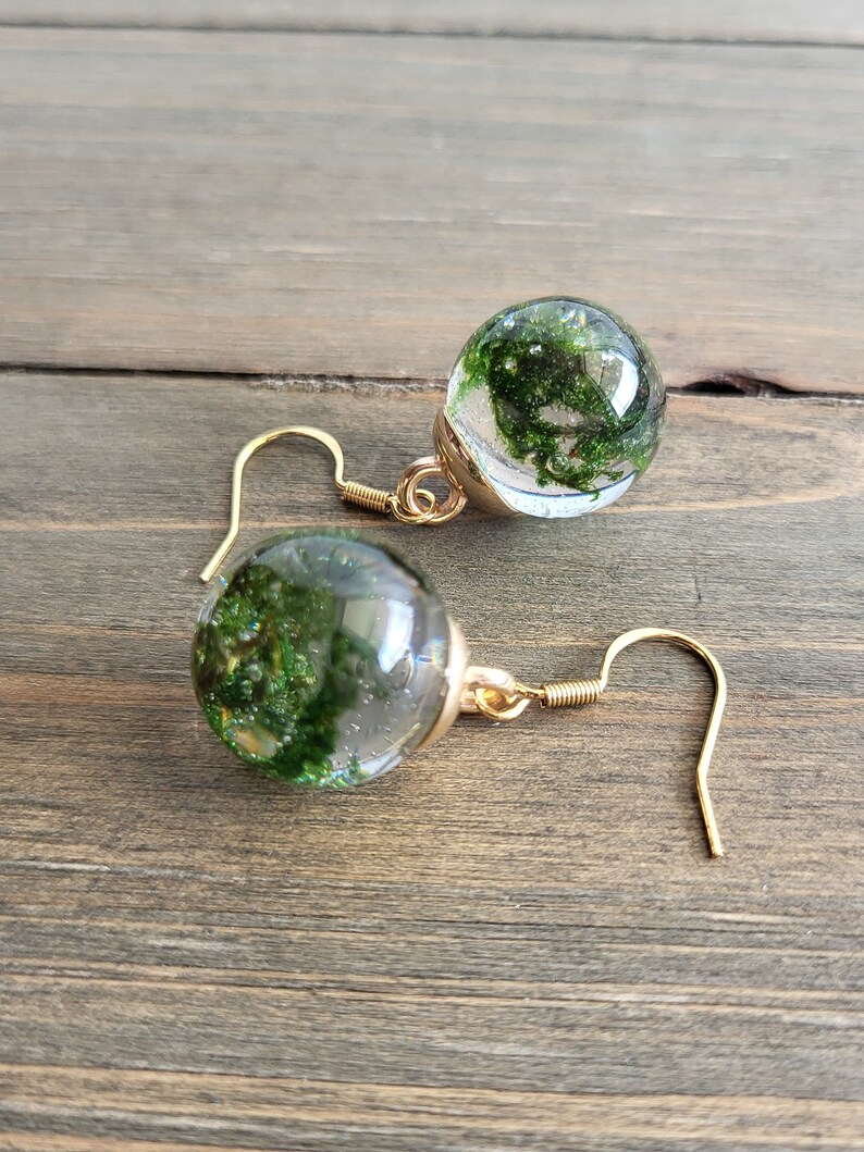 May include: A pair of gold earrings with clear glass orbs containing preserved green moss. The orbs are suspended from gold hooks.