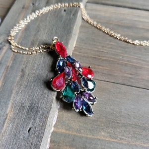 May include: A gold-toned necklace with a pendant featuring an arrangement of colorful, teardrop-shaped gemstones. The gemstones are in shades of red, blue, green, and purple. The necklace is displayed on a weathered wooden surface.