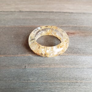Op de afbeelding: Een ring van helder hars met ingebedde gouden vlokken. De ring is dik en heeft een afgeronde vorm.