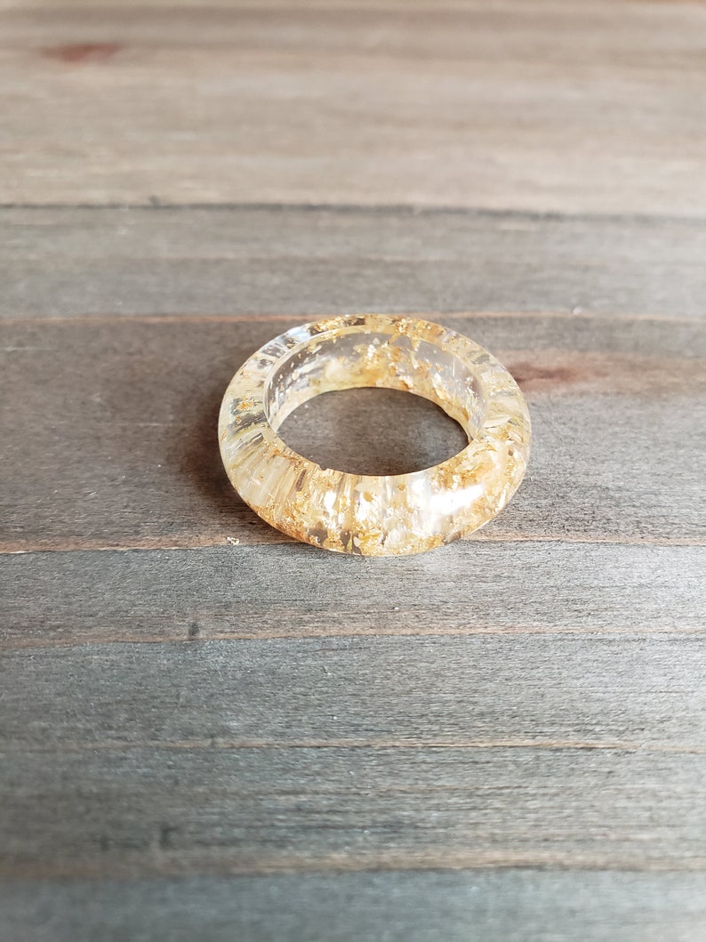 Op de afbeelding: Een ring van helder hars met ingebedde gouden vlokken. De ring is dik en heeft een afgeronde vorm.