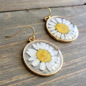 以下が含まれることがあります： 透明な樹脂に封入された本物のデイジーの花が特徴の、金色のイヤリングのペア。 デイジーは白い花びらと黄色の中心部を持っています。 イヤリングには金色のフックが付いています。