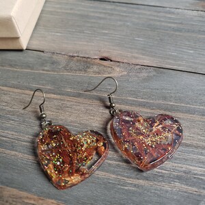 May include: A pair of heart-shaped earrings with gold glitter and brown botanical inclusions encased in clear resin. The earrings have silver hooks.