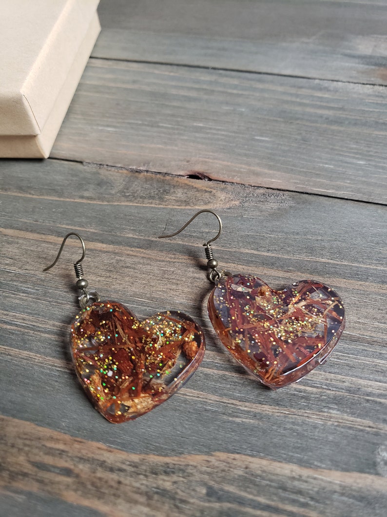 May include: A pair of heart-shaped earrings with gold glitter and brown botanical inclusions encased in clear resin. The earrings have silver hooks.