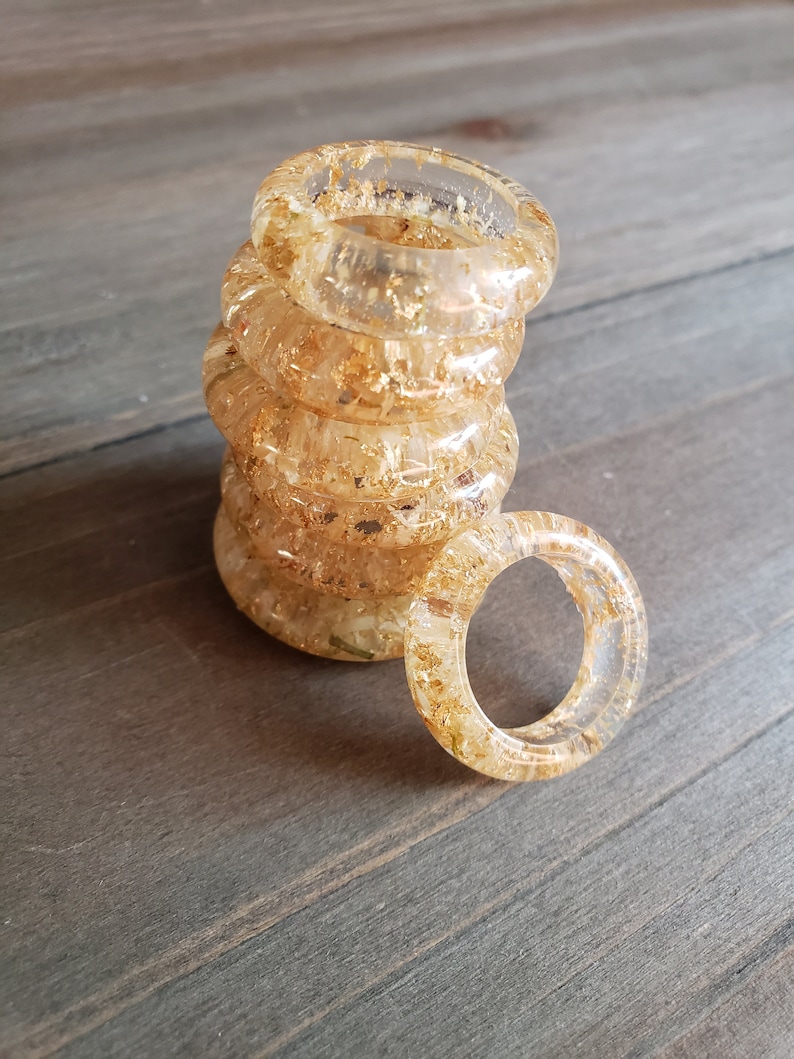 Op de afbeelding: Een stapel van vijf transparante harsringen met gouden vlokken erin. De ringen zijn gestapeld op een houten oppervlak.