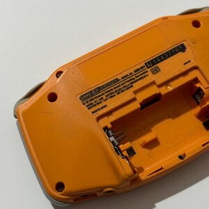 May include: Orange Game Boy Advance handheld gaming console with the back cover removed, revealing the battery compartment and model number AGB-001 AJ 14437765. The console is made in Japan.