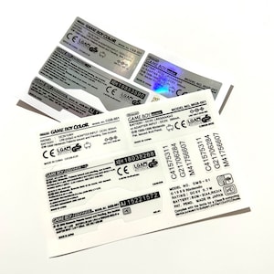 May include: Close-up of a silver and black Game Boy Color sticker with the text "GAME BOY COLOR" and "MODEL NO. CGB-001". The sticker also includes the text "RATING: DC3V 0.8W", "BATTERY: LR6 (AA) or ADAPTER INPUT: DC3V 300mA", "CE", "LGAD", and "MADE IN CHINA CGB-EUR".