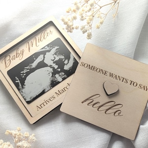 May include: Two wooden plaques. One square plaque features a framed sonogram image with the text "Baby Miller Arrives March". The other square plaque has the text "SOMEONE WANTS TO SAY hello" with a small heart.