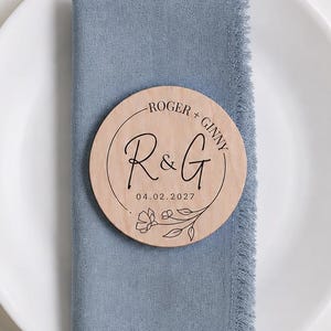 May include: A round wooden place card with black text reading ROGER + GINNY, R & G, and 04.02.2027, featuring a floral line drawing. It rests on a blue linen napkin with frayed edges, placed on a white plate with gold-toned fork, knife, and spoon.