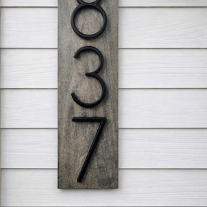 A Vertical modern house number sign featuring the modern black metal numbers "837", Mounted onto a wooden gray staind (aged barrel is the name) rectangular 18 in x 5.5 in x 1/2 in thick plaque. The sign is set against a white exterior wall. Potted plants below on the patio and a small light fixture to the right of the address sign.