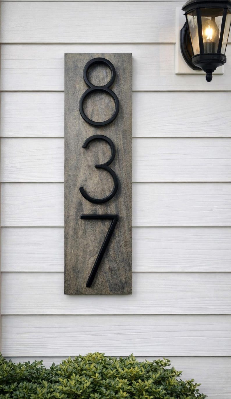 A Vertical modern house number sign featuring the modern black metal numbers "837", Mounted onto a wooden gray staind (aged barrel is the name) rectangular 18 in x 5.5 in x 1/2 in thick plaque. The sign is set against a white exterior wall. Potted plants below on the patio and a small light fixture to the right of the address sign.
