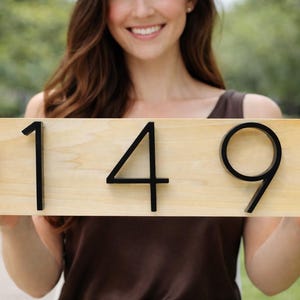 A Horizontal modern house number sign featuring the modern black metal numbers "149", Mounted onto a light wooden rectangular 18 in x 5.5 in x 1/2 in thick plaque. The sign is held up by a women wearing a brown silk top.