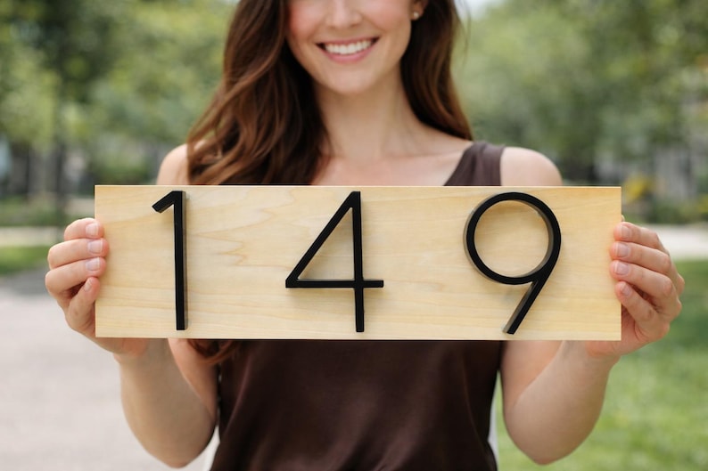 A Horizontal modern house number sign featuring the modern black metal numbers "149", Mounted onto a light wooden rectangular 18 in x 5.5 in x 1/2 in thick plaque. The sign is held up by a women wearing a brown silk top.