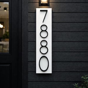 A Vertical modern house number sign featuring the black metal numbers "7880", Mounted onto a wooden white rectangular 24 in x 5.5 in x 1/2 in thick plaque. The sign is set against a dark gray almost black exterior wall, with a black door and a small light fixture above the address sign.