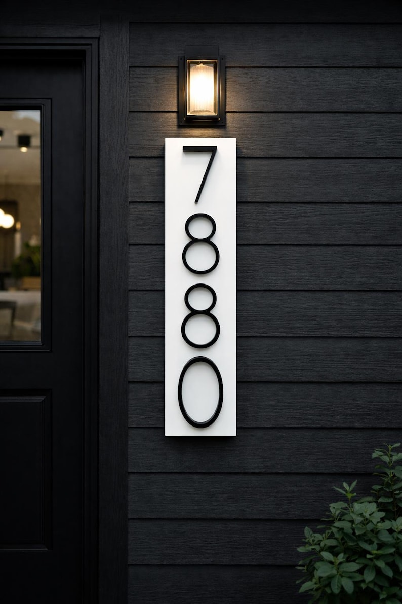 A Vertical modern house number sign featuring the black metal numbers "7880", Mounted onto a wooden white rectangular 24 in x 5.5 in x 1/2 in thick plaque. The sign is set against a dark gray almost black exterior wall, with a black door and a small light fixture above the address sign.