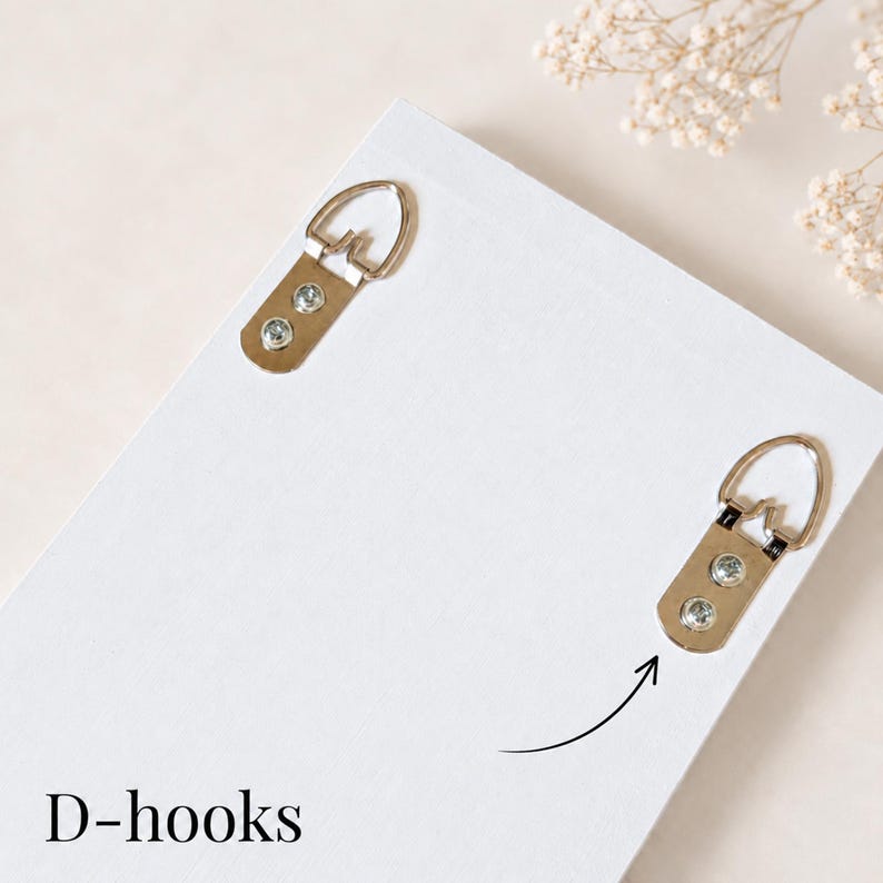 May include: A white rectangular wooden board featuring two silver metal D-hooks attached with screws. The D-hooks are positioned near the top corners of the board, designed for wall mounting. The text D-hooks is printed in black at the bottom.