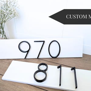 White Horizontal 18 in x 5.5 in x 1/2 in thick rectangular address plaques with modern black metal numbers. One plaque displays the numbers 970, and the other shows 811. The words "CUSTOM MADE" are in a black arrow shape in the background.