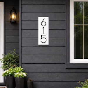 A Vertical modern house number sign featuring the modern black metal numbers "615", Mounted onto a wooden white rectangular 18 in x 5.5 in x 1/2 in thick plaque. The sign is set against a gray exterior wall. Potted plants below on the patio to the left of the address sign and a small light fixture next to the front door.