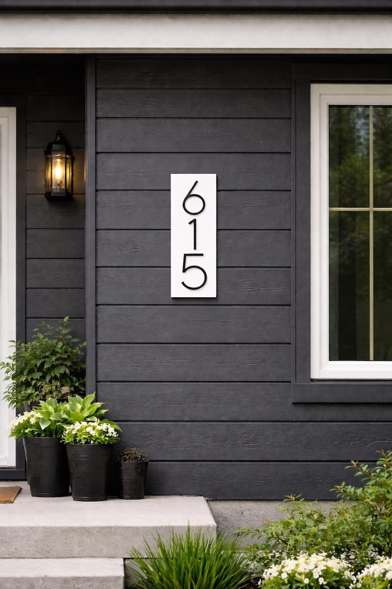 A Vertical modern house number sign featuring the modern black metal numbers "615", Mounted onto a wooden white rectangular 18 in x 5.5 in x 1/2 in thick plaque. The sign is set against a gray exterior wall. Potted plants below on the patio to the left of the address sign and a small light fixture next to the front door.