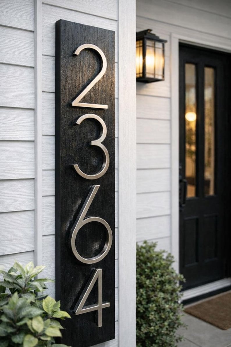 A Vertical modern house number sign featuring the modern silver metal numbers "2364", Mounted onto a wooden black rectangular 24 in x 5.5 in x 1/2 in thick plaque. The sign is set against a white exterior wall, with a black door and a small light fixture next to the front door.