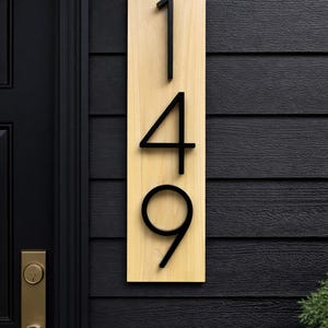 A Vertical modern house number sign featuring the modern black metal numbers "149", Mounted onto a light tone wooden rectangular 18 in x 5.5 in x 1/2 in thick plaque. The sign is set against a dark gray exterior wall. Potted plants below on the patio to the right of the address sign and a black front door with brass handle to the left.